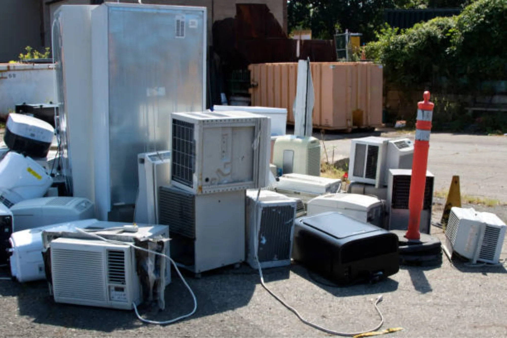 Scrap yard collection of old outdoor air conditioner units and refrigerators stacked for metal recovery and safe gas dismantling.